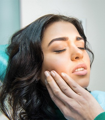 Woman with dark hair in dental chair holding hand to jaw in pain