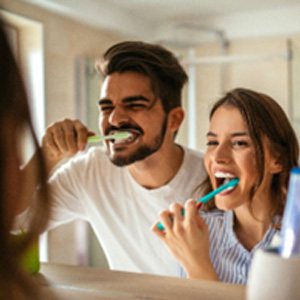 Man and woman smiling while brushing their teeth in bathroom