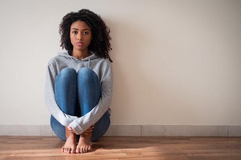 a young female wearing a gray hoodie and jeans sitting against a wall and looking fearful and stressed
