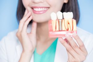 Dentist smiling in the background holding her jaw while holding out a model dental implant to the foreground with her other hand